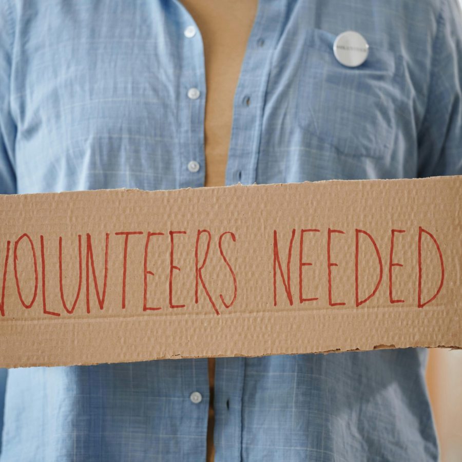 Person holding a sign reading 'Volunteers Needed' to encourage community support.